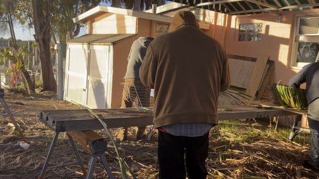 Three crew members weaving nest baskets at a workbench under the canopy